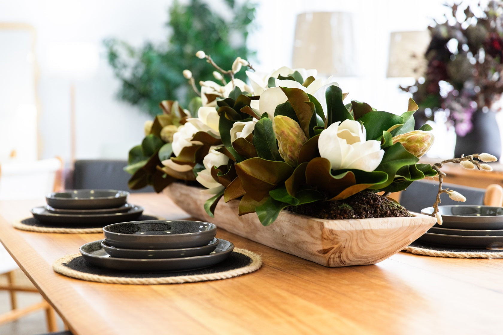 Decorative floral arrangement in a wooden bowl on a table with black plates and placemats.